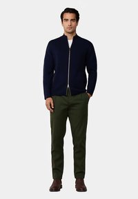 Marinblå zip-up tröja i slät tyg, kombinerad med olivgröna byxor med rak skärning och bruna ankelstövlar.