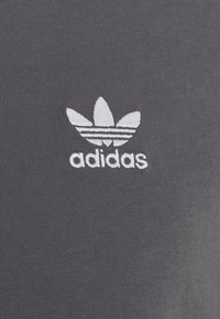 Grå bomullstyg med en vit broderad Adidas trefoil-logotyp ovanför varumärkesnamnet i ett rent sans-serif typsnitt.