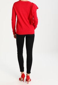 Femme portant un pull rouge à manches volantées, un pantalon noir moulant et des chaussures à talons hauts rouges, debout de dos sur fond blanc.