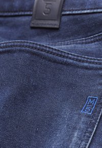 Tissu en denim bleu foncé, avec une étiquette en cuir portant le chiffre "5", des coutures bleues et une texture lisse, mettant en valeur les détails des coutures.