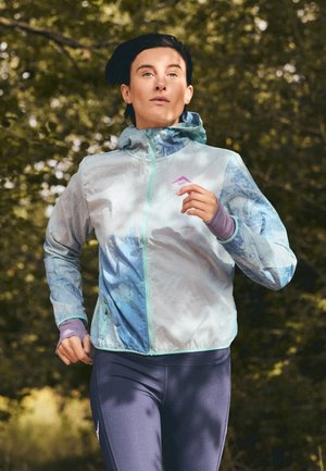 Persona corriendo al aire libre con una chaqueta ligera de color azul claro con estampado y leggings deportivos morados en un parque con hojas.