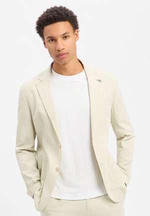 Junger Mann mit lockigem Haar trägt eine beige Anzugjacke über einem weißen Hemd, steht vor einem schlichten weißen Hintergrund mit in die Taschen gesteckten Händen.