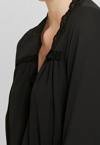 Svart blus med delikat svart spetskant längs halsringningen, i mjukt, flödande tyg med plisserad design.