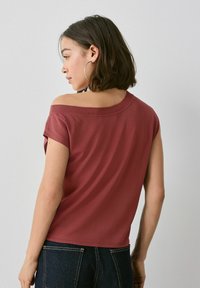 Camiseta burdeos de hombros descubiertos con mangas cortas, textura suave, corte relajado y detalle de costura en contraste, combinada con jeans de mezclilla oscura.