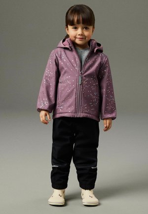 Copil tânăr în picioare, purtând o jachetă mov cu glugă și pete argintii, pantaloni negri cu zone reflectorizante și cizme deschise la culoare cu modele.