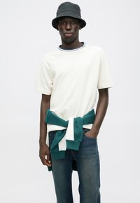 Κρέμα κοντομάνικη μπλούζα με μπλε ριγέ γιακά, συνδυασμένη με μπλε τζην. Ένα πράσινο και λευκό φούτερ δεμένο στη μέση. Μαύρο καπέλο bucket.