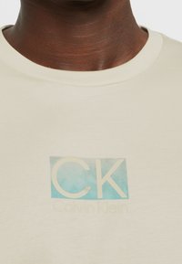 Close-up of a person wearing a cream t-shirt with "CK" in large letters and "Calvin Klein" printed below on a blue square background.