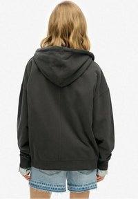 Sudadera gris oscuro con una gran capucha, corte holgado y puños acanalados. Combinada con pantalones cortos de mezclilla con dobladillos deshilachados y una camisa de rayas en capas.