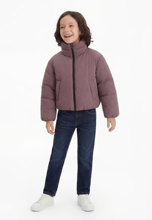 Bērns ar cirtainiem matiem, valkājot violeti rozā pūkainu jaku, tumši zilas džinsas un baltas sporta kurpes, stāv un smaida pret baltu fonu.