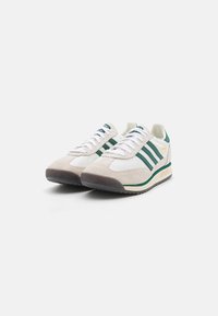 Zapatillas Adidas SL 72 en blanco y beige con rayas y acentos verdes, cordones blancos y suelas texturizadas, mostradas en un ángulo sobre un fondo blanco.