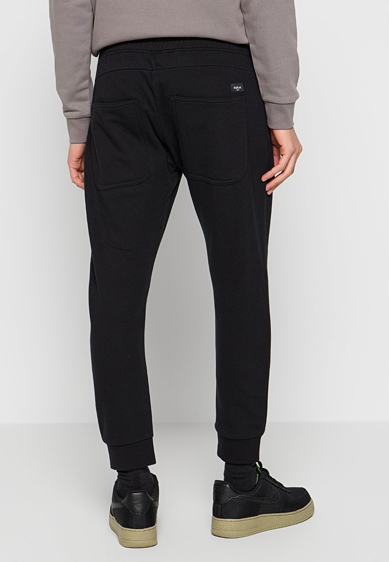 Pantaloni jogger neri con caviglie a costina, due tasche posteriori e un'etichetta in vita, abbinati a scarpe nere e calzini scuri.