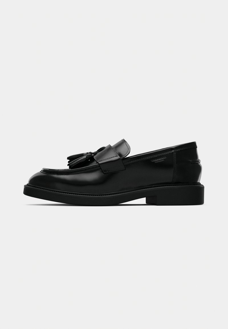 Mocassins en cuir noir avec un détail de couture surélevé, semelle en caoutchouc plate et accents décoratifs de glands à l'avant. Texture lisse.