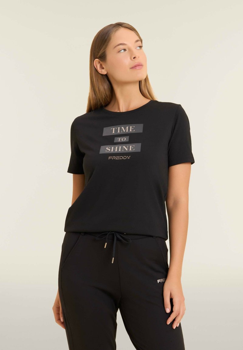 Camiseta negra de manga corta con texto plateado "HORA DE BRILLAR" y acentos de strass. Tejido suave, corte relajado, combinada con pantalones negros.