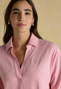 Pikkade tumedate juustega naine kannab kuldseid rõngaste kõrvarõngaid ja roosat kraejope, millel on tagasihoidlik kirjaõmblus, beeži taustal.