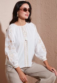 Blusa bianca con ricami floreali dorati, vestibilità ampia, scollo a stringere e maniche a sbuffo; indossata con pantaloni in lino chiari e occhiali da sole.