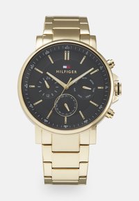 Zelta un melna pulksteņi ar apaļu seju, kas ir aprīkots ar vairākiem ciparnīcām, strukturētu melnu fona un zelta saiti. Uzraksts "HILFIGER".