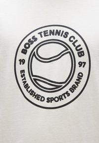 Marškinėliai su juodu apskritu spaudiniu, kuriame yra užrašas „BOSS TENNIS CLUB“, teniso kamuoliuko grafika ir užrašas „ESTABLISHED SPORTS BRAND“ ant kreminio fono.