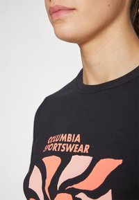 Melna T-kreka ar apaļu kakla izgriezumu, kas rotāta ar krāsainu spirāles dizainu rozā un gaiši persiku krāsā, kā arī "Columbia Sportswear" logotipu.