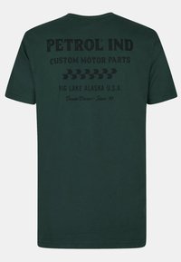 Zelené bavlněné tričko s černým potiskem na zádech s nápisem "PETROL IND," vlastním textem a motivy šipky. Krátký rukáv a kulatý výstřih.