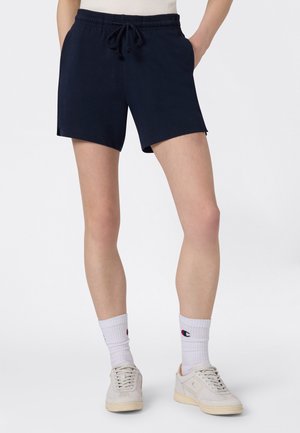 Persona che indossa pantaloncini blu navy con elastico in vita e coulisse, calzini bianchi al polpaccio con logo e scarpe da ginnastica bianche, in piedi su uno sfondo uniforme.