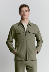 Olijfgroene ritsjack met twee borstzakken, lange mouwen en een kraag. Gladde stof met een gestructureerd ontwerp, gecombineerd met bijpassende broek.