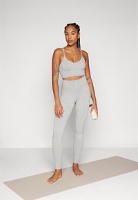 Sivi atletski crop top i tajice s visokim strukom. Glatka tkanina, uski kroj, s tankim naramenicama. Stojeći na prostirci, drži bocu vode.