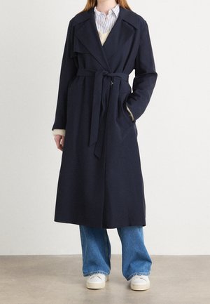 Personne portant un long manteau ceinturé bleu marine foncé par-dessus un jean bleu et des baskets blanches, debout devant un fond clair uni.