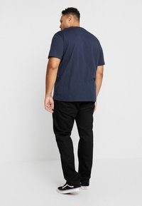 Hombre vestido con una camiseta azul marino, pantalones negros y zapatillas en blanco y negro, de pie de espaldas contra un fondo gris claro y liso.
