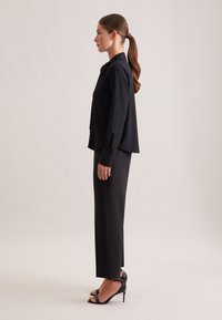 Blusa nera e pantaloni neri su misura. La blusa presenta un colletto e maniche lunghe. I pantaloni hanno un taglio dritto e una sottile lucentezza. Tacchi neri.