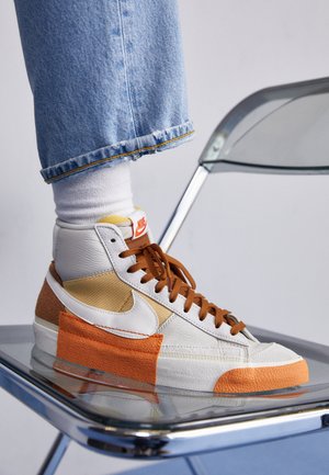 Person mit weißen Socken, blauen Jeans und weißen, orangefarbenen und braunen Nike High-Top-Sneakern, die den Fuß auf einen transparenten Plastikstuhl legen.