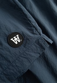 Tessuto blu navy con una superficie texturizzata, caratterizzato da una toppa circolare nera con un design stilizzato della lettera bianca, con dettagli di cucitura.