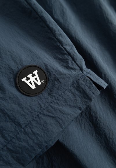 Tissu bleu marine avec une surface texturée, présentant un patch circulaire noir avec un design de lettres stylisées blanches, détail de couture sur les coutures.