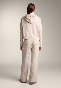 Pletená mikina s kapucí a kalhoty s širokými nohavicemi v světle béžové barvě, s výrazným copánkovým vzorem, žebrovanými manžetami a volným střihem.