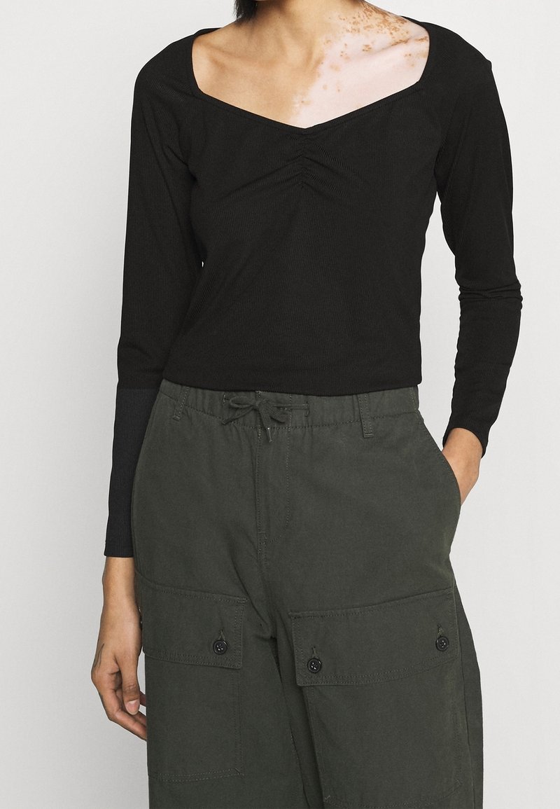 Svart långärmad topp med fyrkantig halsringning, med subtila rynkor i mitten. Kombineras med olivgröna cargobyxor med fickor.
