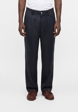 Homme portant un pantalon plissé bleu marine foncé, un t-shirt blanc et des chaussures en cuir marron à lacets, debout devant un fond clair uni.