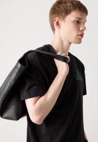Svart lær tote bag som henger over skulderen, med en glatt tekstur og minimalisk design. Mannen har på seg en svart T-skjorte med grønt logo-detail.
