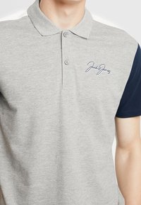 Šedé polo tričko s límečkem, krátkými námořnickými modrými rukávy, dvěma knoflíky a vyšitým logem "Jack & Jones" v námořnické barvě. Měkká texturovaná látka.