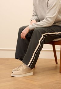 Beige gestrickte Sneaker mit strukturiertem Obermaterial, flacher weißer Sohle, getragen mit einem grauen Sweatshirt und dunklen grünen Hosen mit cremefarbenen Seitenstreifen.