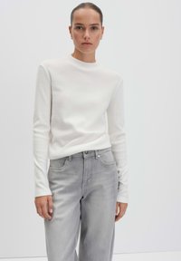 Weißer Pullover mit langen Ärmeln aus weichem Stoff, kombiniert mit hellgrauen, lässigen Jeans mit weitem Bein, die über Taschen und einen Knopfverschluss verfügen, minimales Design.