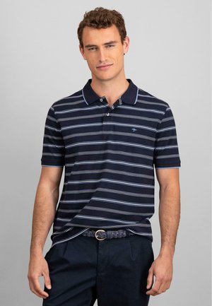 Marineblauwe polo met witte en lichtblauwe strepen, korte mouwen, een kraag en een placket met twee knopen. Gestyled met marineblauwe broek en een gevlochten riem.