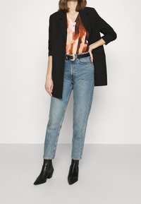 Blazer nero sopra una blusa astratta multicolore, abbinato a jeans a vita alta azzurri chiari e stivaletti neri a punta.