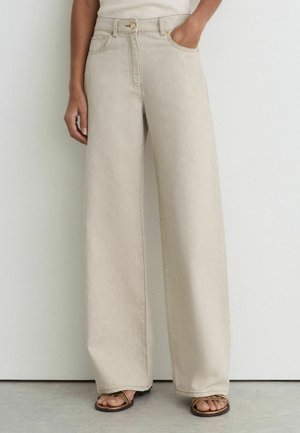 Kvinde iført beige jeans med høj talje og vide ben med lommer foran og bagpå, kombineret med brune remsandaler, står mod en hvid væg.