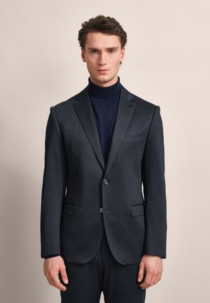 Dunkelgrauer maßgeschneiderter Blazer aus einem subtil strukturierten Stoff. Verfügt über zwei Knöpfe, Reverskragen und seitliche Taschen. Trägt man über einem marineblauen Rollkragenpullover.