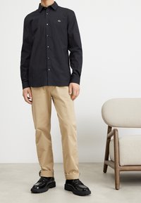 Camisa negra de manga larga con botones y logo bordado, pantalones beige y zapatos negros con suelas gruesas, sobre un fondo neutro.