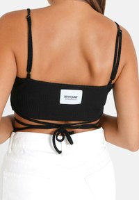 Top corto nero a costine con spalline sottili e regolabili, caratterizzato da un dettaglio a nodo sul retro e un'etichetta bianca con logo. Abbinato a jeans bianchi.