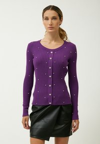 Cardigan viola a costine con maniche lunghe, impreziosito da bottoni decorativi in argento e accenti di gemme trasparenti, abbinato a una gonna di pelle nera.