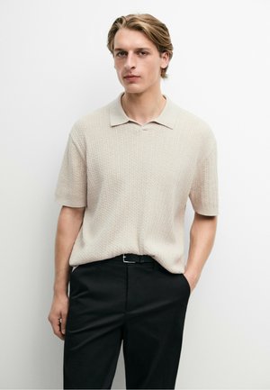 Junger Mann mit blondem Haar, der ein beiges, kurzärmeliges, gestricktes Poloshirt und schwarze Hose trägt, steht vor einer schlichten weißen Wand.