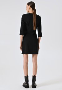 Schwarzes Kleid mit Dreiviertelärmeln, tailliertem Schnitt und ausgestellt Saum. Kombiniert mit kniehohen schwarzen Lederstiefeln mit dicken Sohlen.