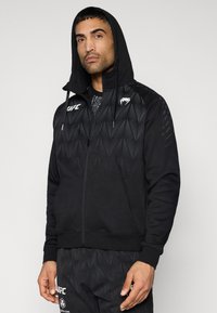 Hoodie noir à fermeture éclair avec un motif géométrique texturé. Il présente une capuche doublée, des poignets élastiques et un petit logo sur la poitrine.