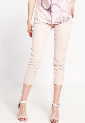 Femme portant un pantalon court rose pâle et un chemisier à fleurs rose clair, debout avec une main sur la hanche, portant des talons beiges à brides à la cheville.